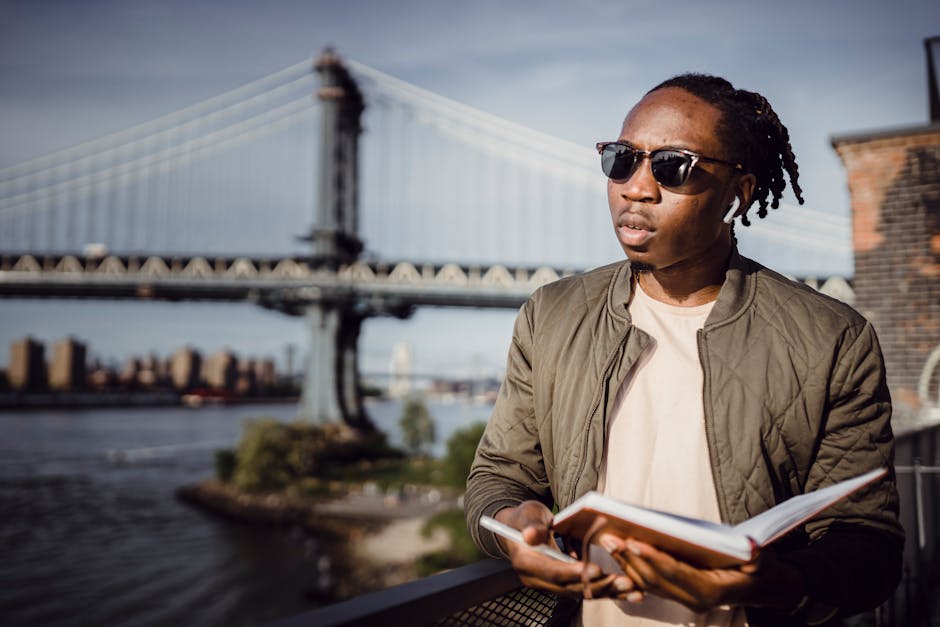 A stylish young man in sunglasses stands by the Brooklyn Bridge, enjoying the sunny day with a notebook.